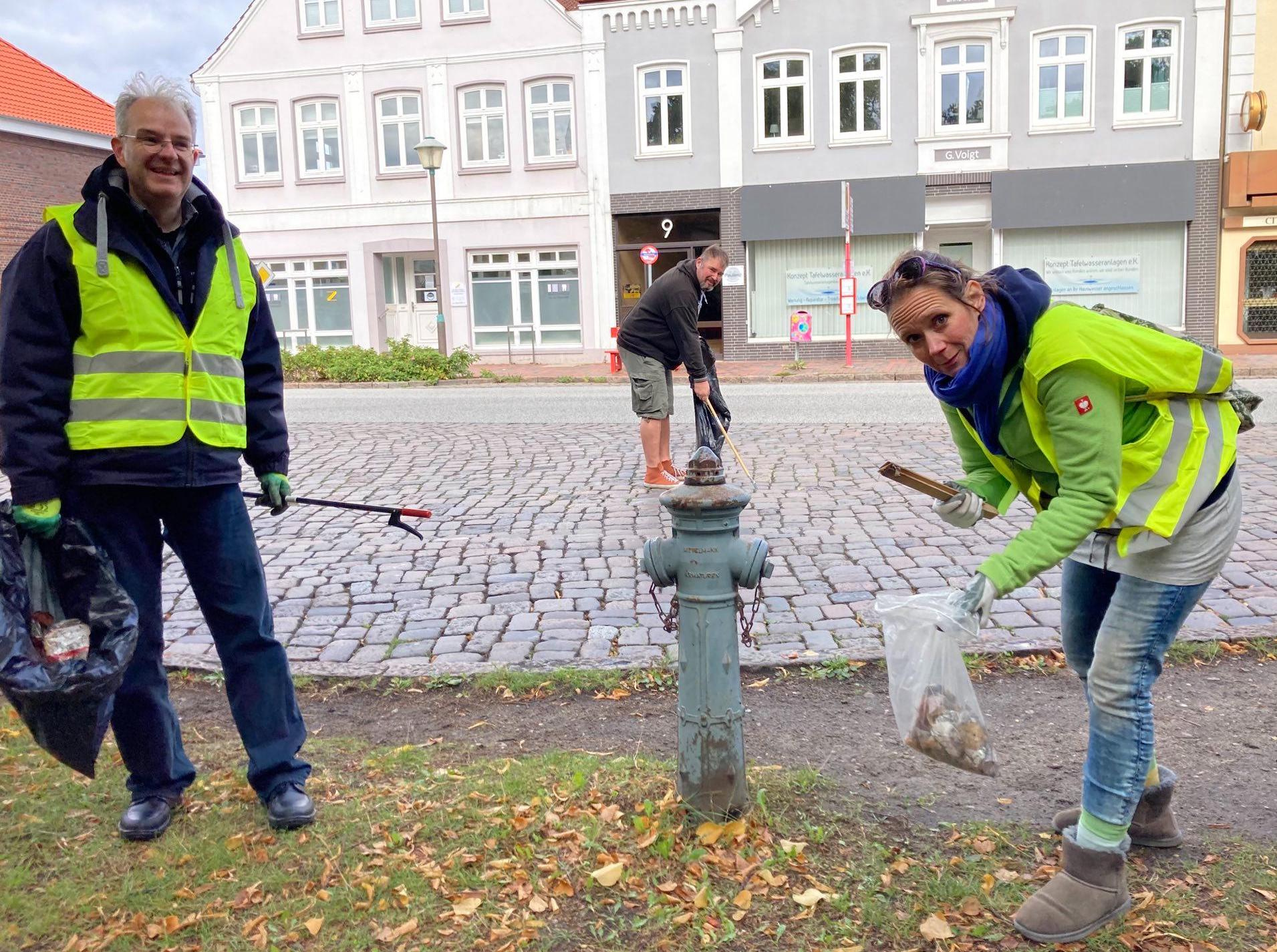 Seitliches Foto von drei Aktiven der Uetersener Grünen beim Müll sammeln. Sie tragen gelbe Warnwesten und Schutzhandschuhe und lächeln in die Kamera. In den Händen halten sie Greifer, mit denen sie Abfall in verschiedene Müllsäcke werfen. Im Hintergrund sieht man eine gepflasterte Straße und einige Häuser.